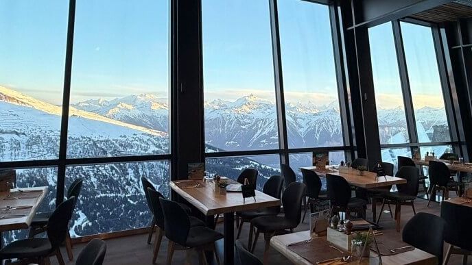 Gedeckte Tische im Panoramarestaurant mit Blick durch grosse Fenster auf verschneite Berge.