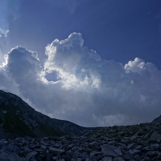 Hoch aufragende Quellwolken über einem felsigen Berghang unter blauem Himmel.