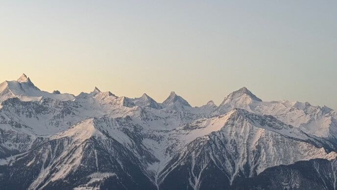 Verschneite Gipfel der Walliser Alpen im warmen Licht über dunklen Berghängen.